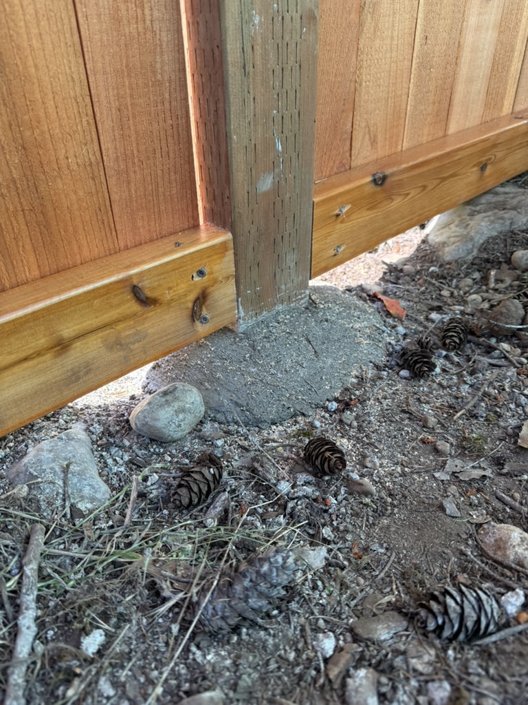 Close-up of a domed, above-grade concrete footing around a pressure-treated fence post